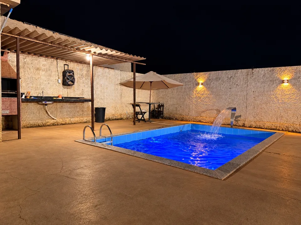 Piscina iluminada à noite, com a churrasqueira coberta ao fundo